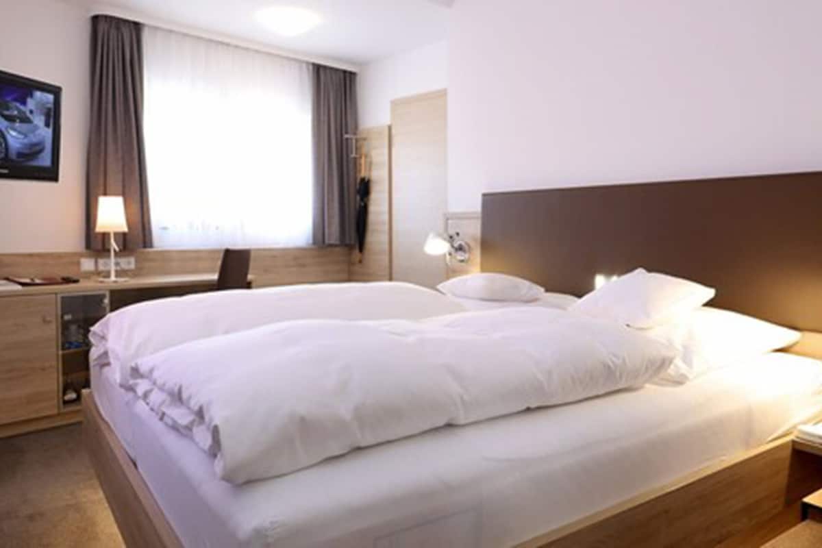 A hotel room with a large bed, a desk with a computer, a TV mounted on the wall, and a window with curtains.