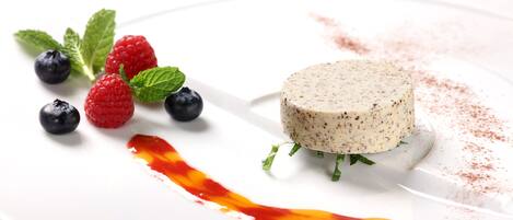 A dessert plate with a round, speckled dessert, raspberries, blueberries, mint leaves, and a red sauce.