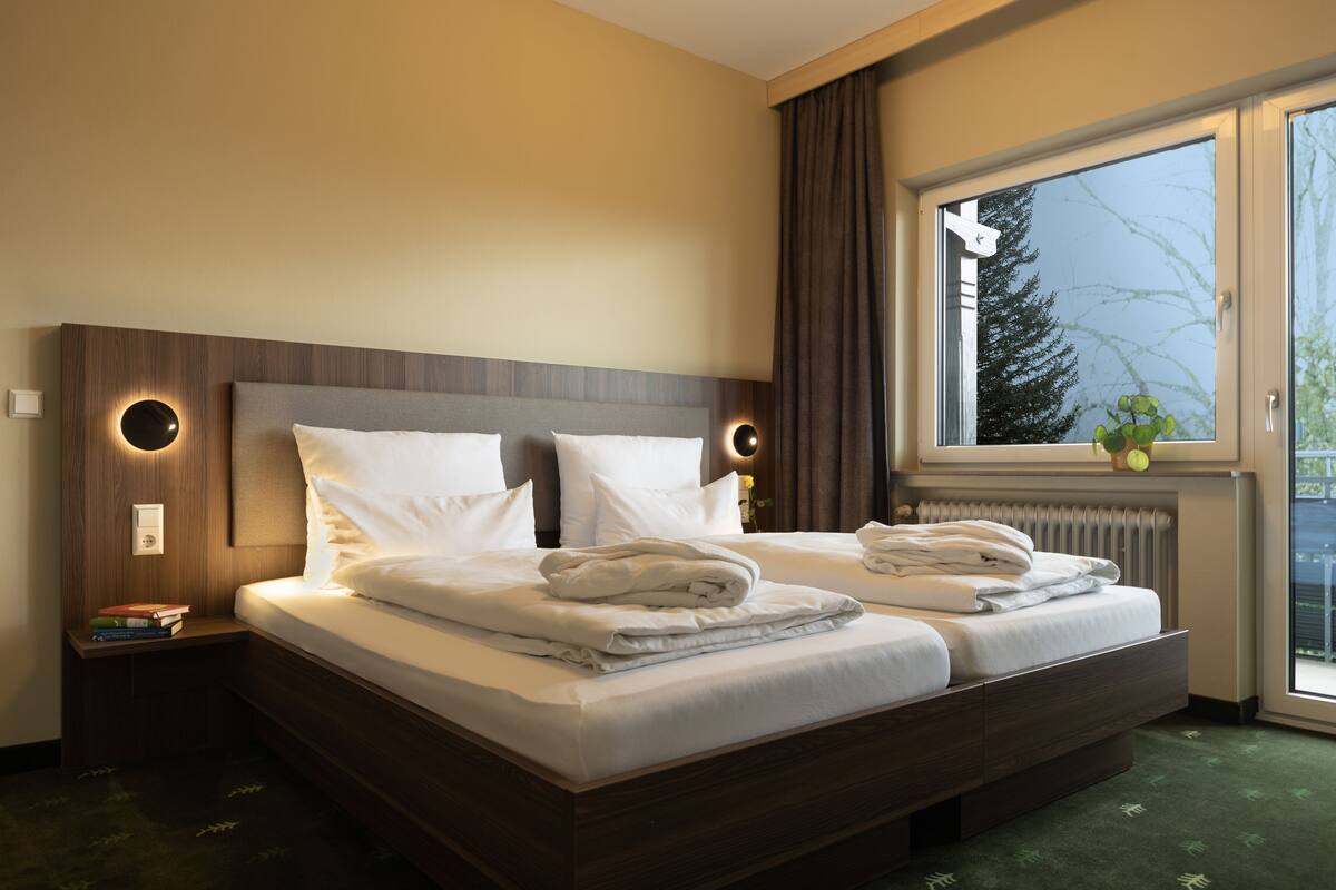 A double bed with white linens, a wooden headboard, bedside tables with lamps, and a window with a view of trees.
