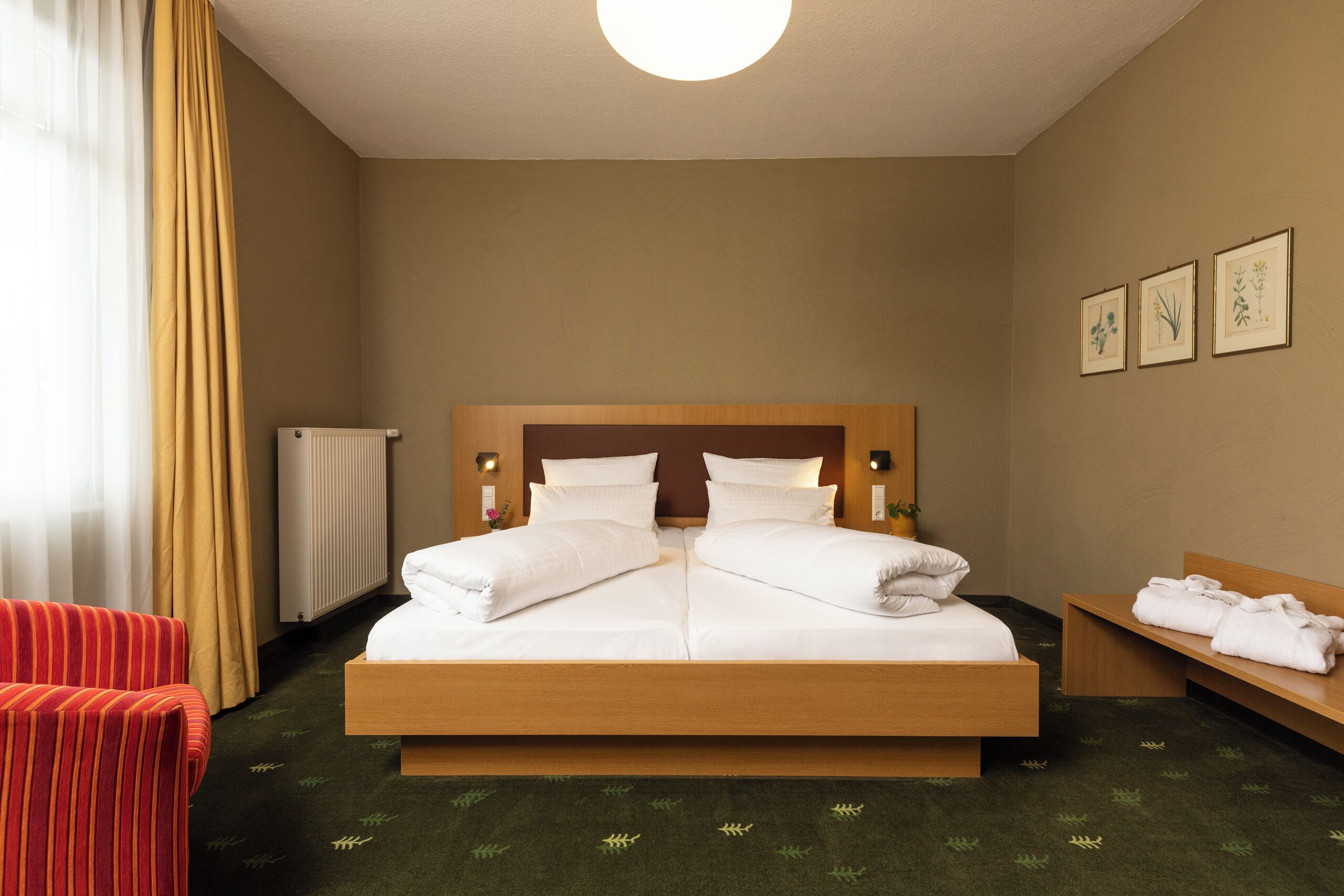 Habitación de hotel con una cama grande, cabecera de madera, mesita de noche con toallas, una silla roja, radiador e impresiones botánicas enmarcadas en la pared.