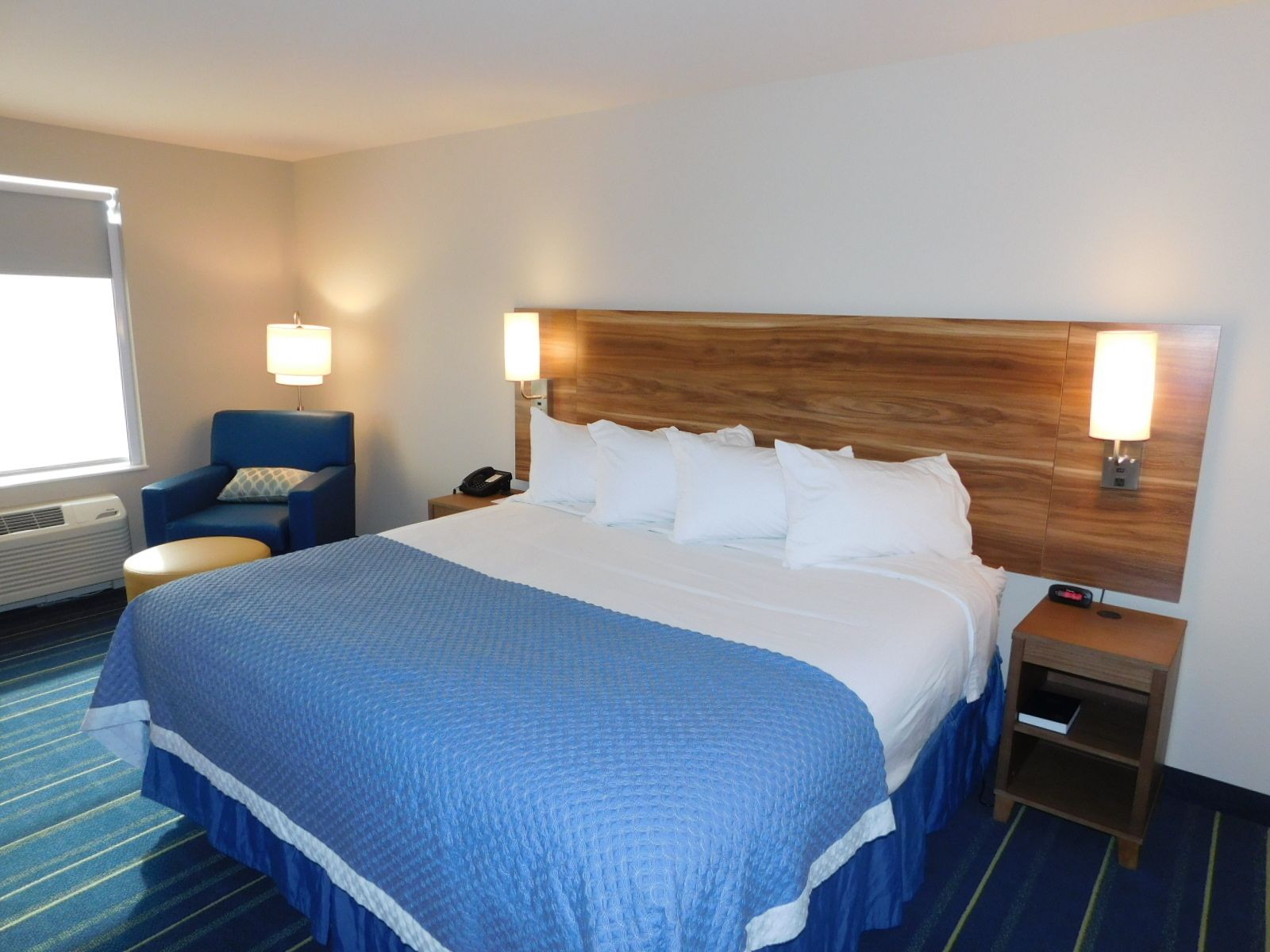 A hotel room with a large bed, a blue chair, a bedside table with a lamp, and a wall-mounted telephone.