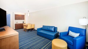 Habitación de hotel con sofá azul, una mesita redonda pequeña, televisión y una kitchenette al fondo.