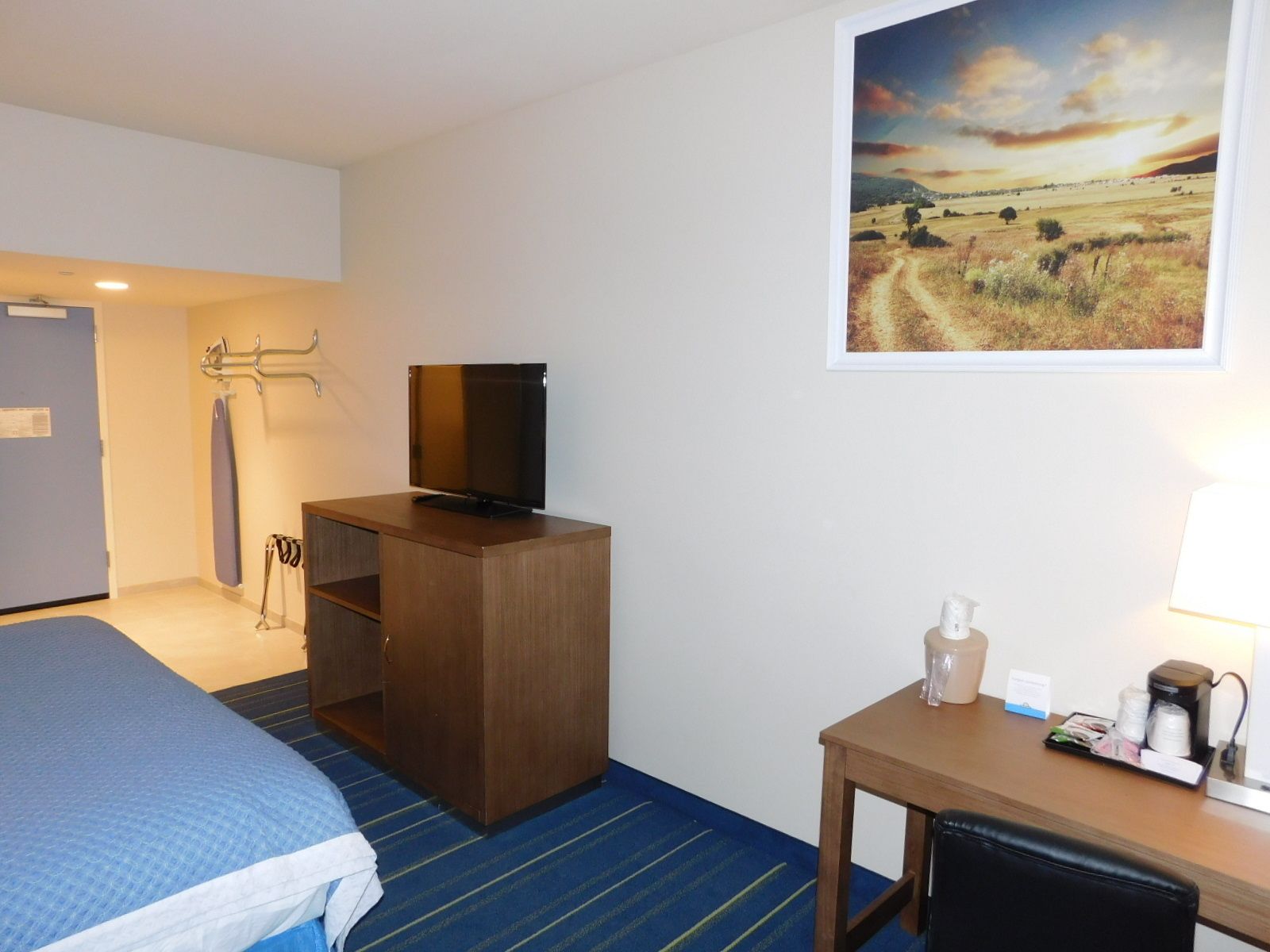 Une chambre d’hôtel avec un lit, un bureau doté d’une lampe, une télévision et un tableau représentant un paysage.