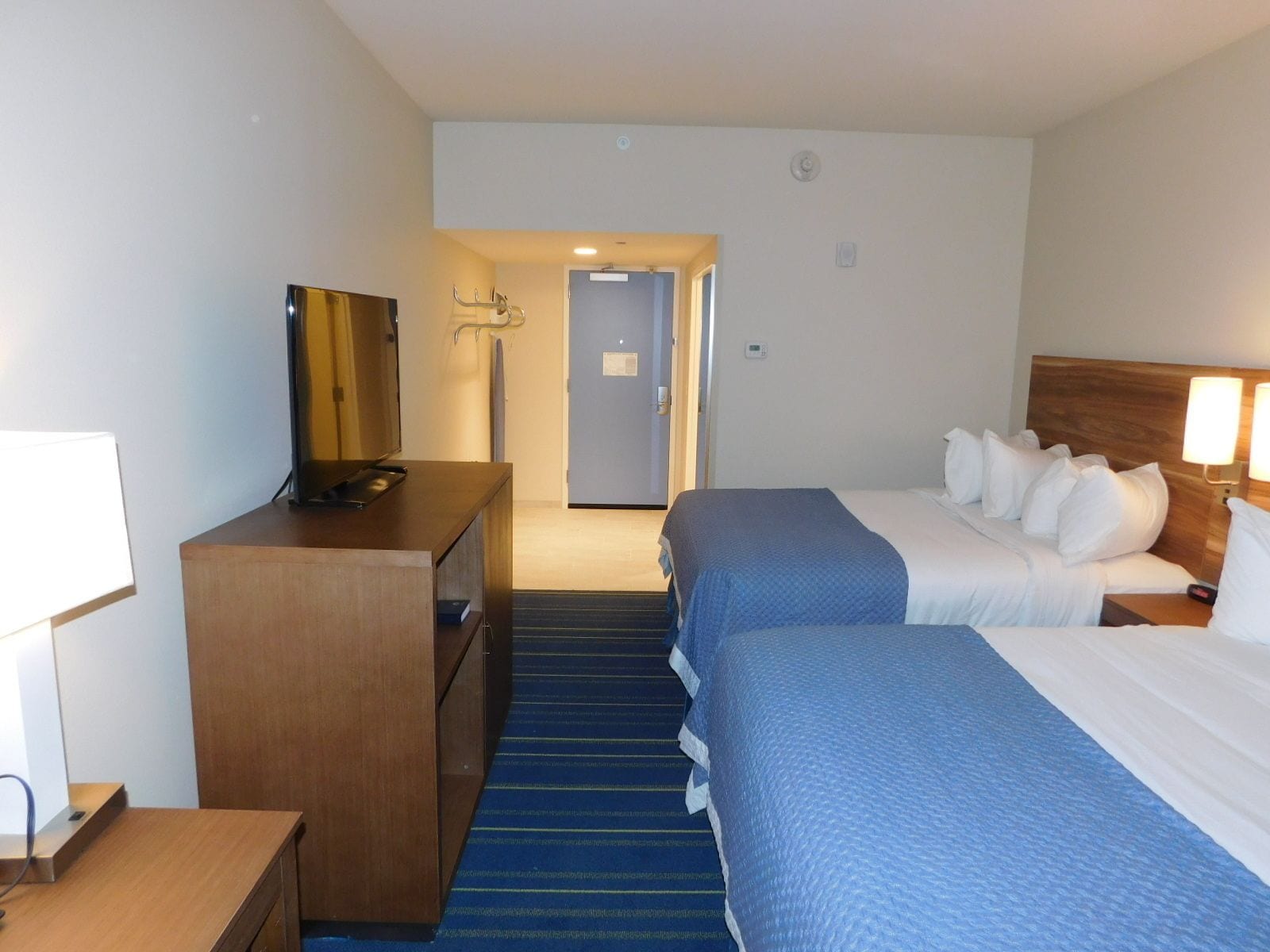 Una habitación de hotel con escritorio, televisor, cama con ropa de cama azul y cabecero con lámpara.
