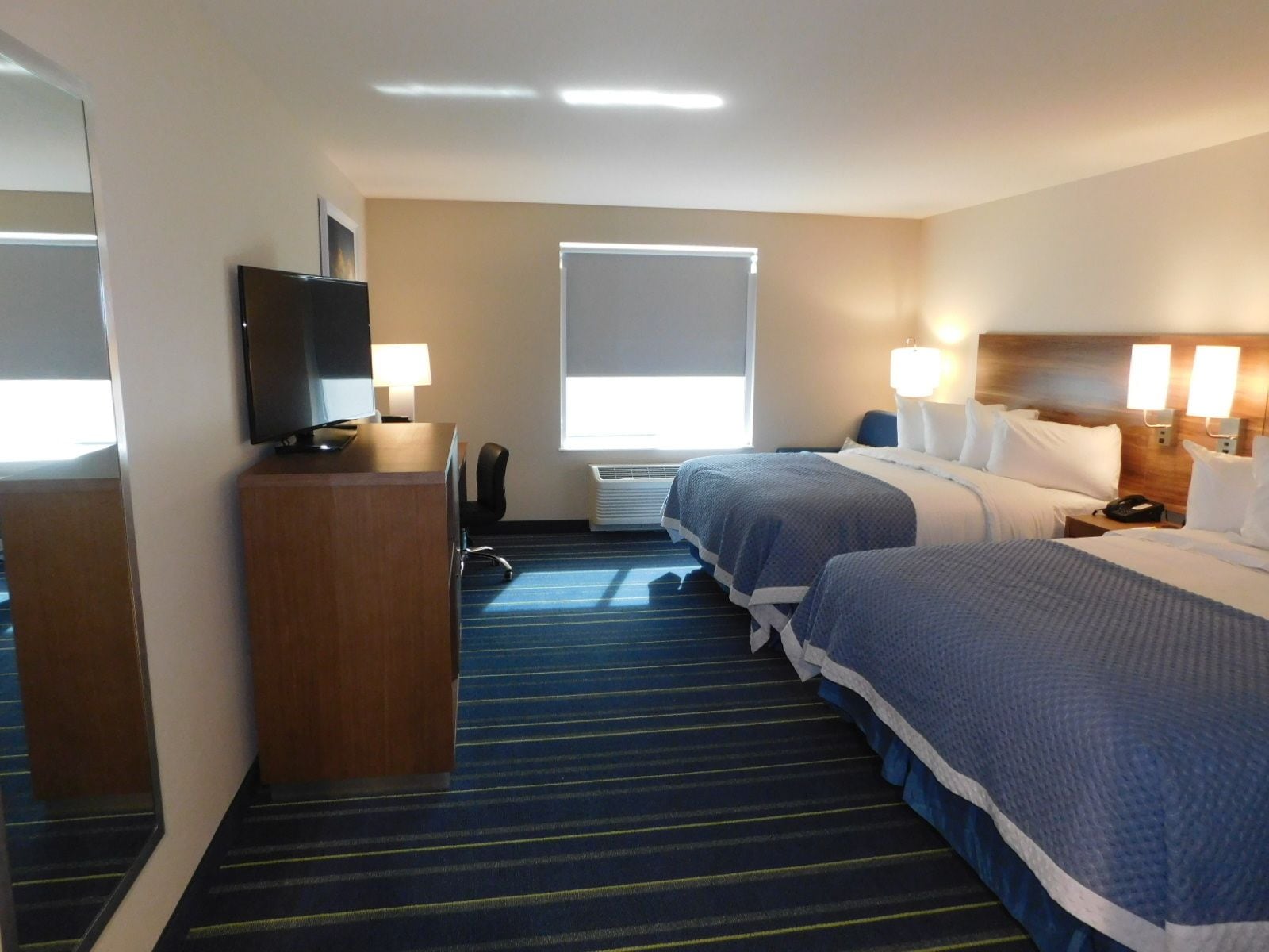 Une chambre d’hôtel avec deux lits, un bureau avec une télévision, une chaise et une fenêtre avec des stores.