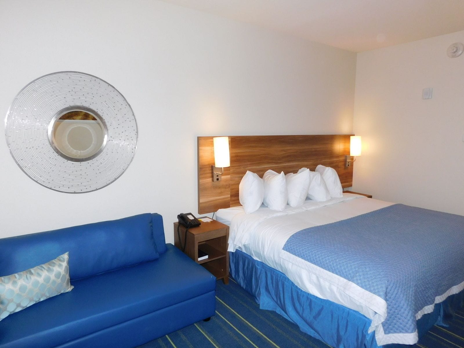 A hotel room with a bed, a blue sofa, a bedside table with a phone, and a wall-mounted light fixture.
