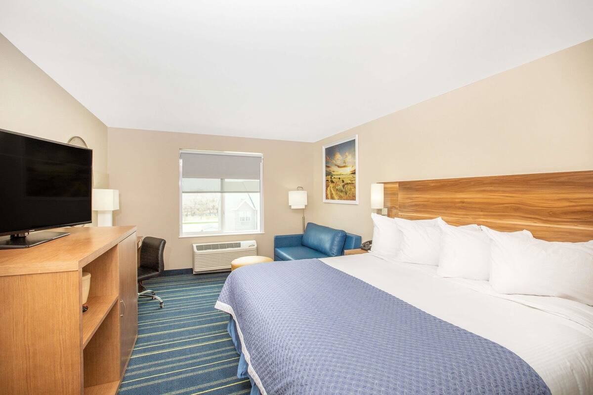 A hotel room with a large bed, a desk with a television, a blue chair, and a window with blinds.