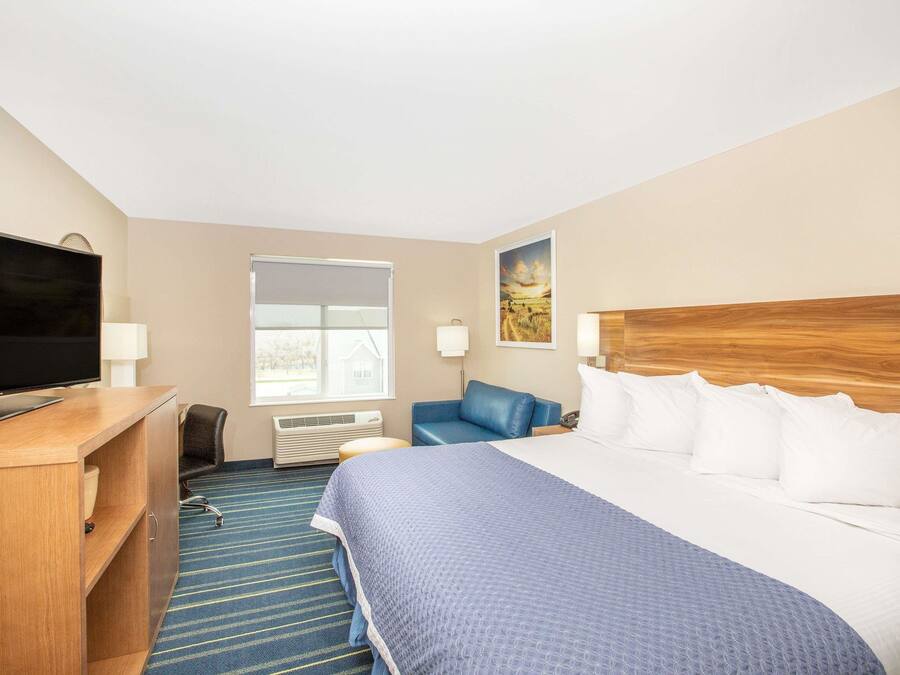 A hotel room with a large bed, a desk with a television, a blue chair, and a window with blinds.