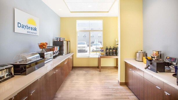 Un área de desayuno con un cartel que dice 'Daybreak Breakfast', un bufé con diversas opciones de comida, una máquina de café y una ventana con vistas al exterior.