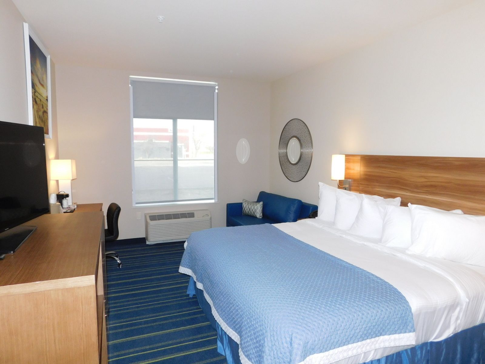 A hotel room with a large bed, a desk with a TV, a window, and a wall-mounted mirror.