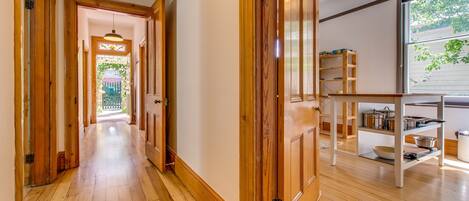 A hallway with wooden floors and doors, leading to a bright room with a window and a shelf with kitchen items.