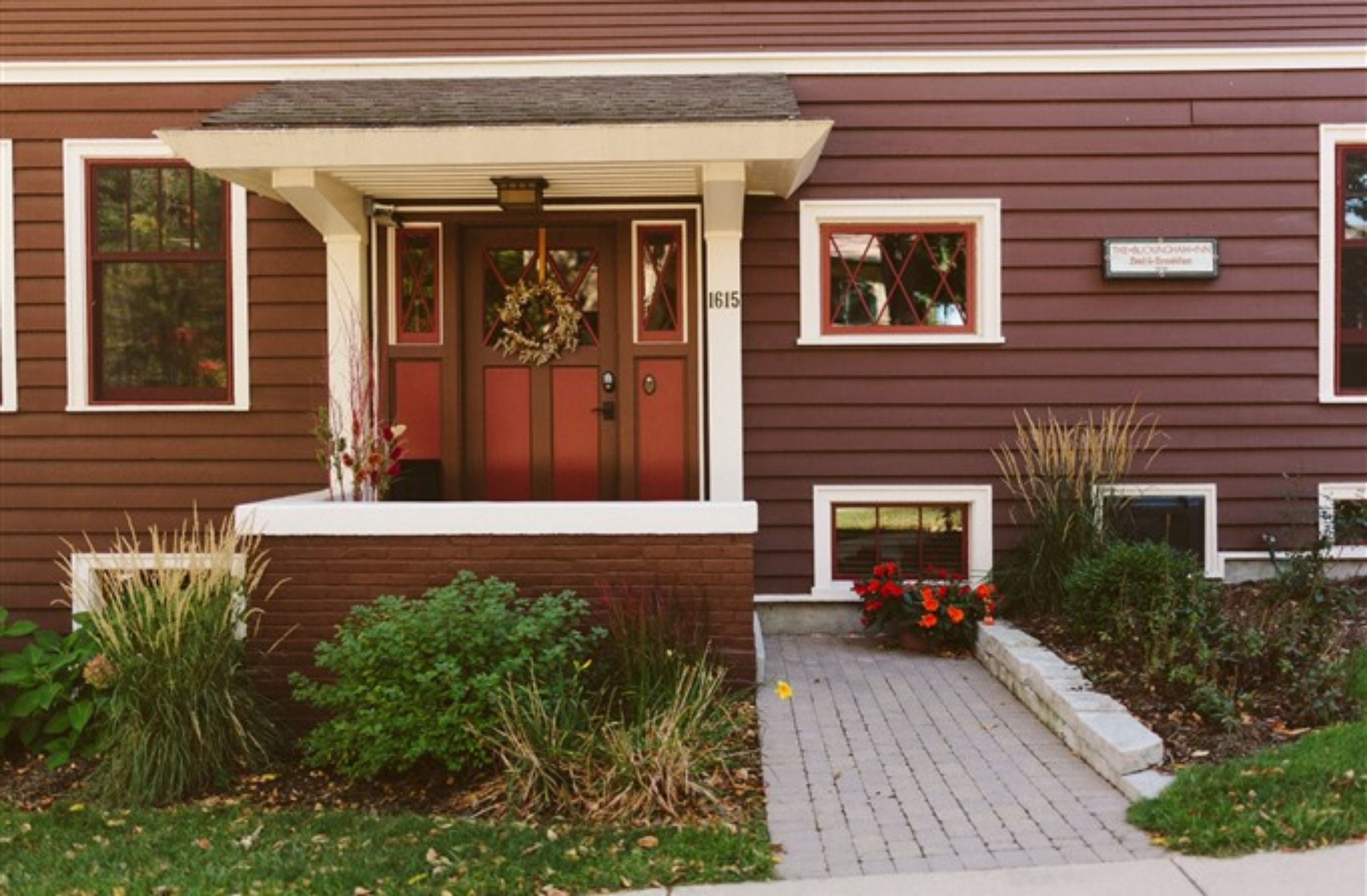 A two-story house with a front porch, a red door, and a small garden area with red flowers and plants.