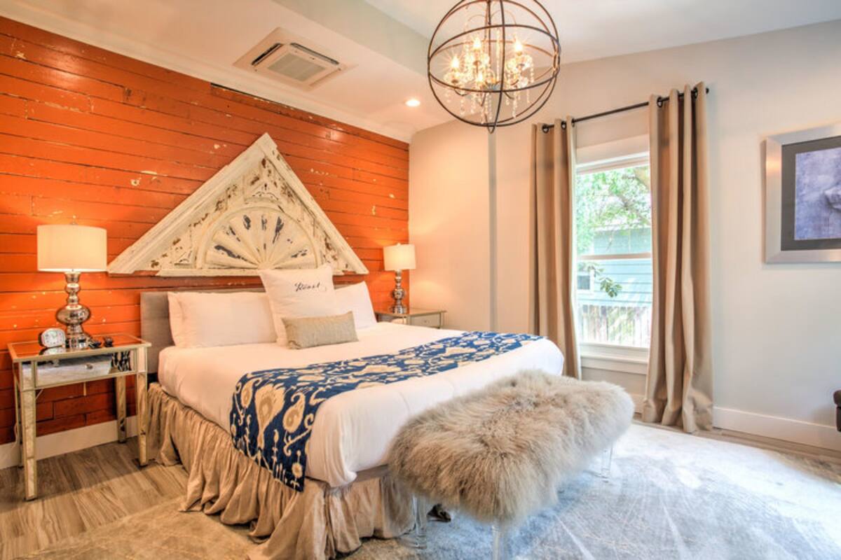 A bedroom with a wooden headboard, a large bed with a patterned bedspread, two bedside tables with lamps, a chandelier, and a view of the outdoors through a window with curtains.