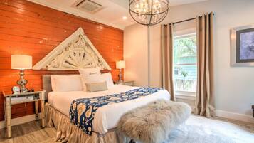 A bedroom with a wooden headboard, a large bed with a patterned bedspread, two bedside tables with lamps, a chandelier, and a view of the outdoors through a window with curtains.
