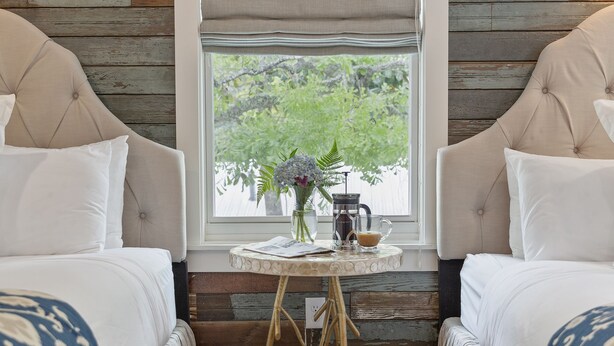 A bedroom with two beds, a window with a view of greenery, a small table with a vase and a coffee pot, and a wooden floor.