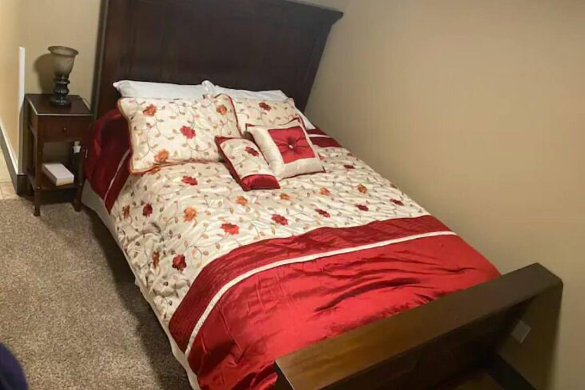 Un lit bien fait, avec une couette à motifs floraux et des touches de rouge, une tête de lit en bois et une table de chevet avec une lampe.