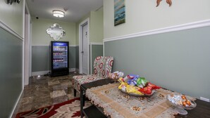 Un couloir avec un distributeur automatique, une chaise à motifs, un plateau rempli de collations et une horloge au mur.