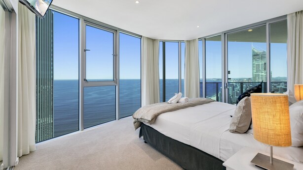 A modern hotel room with a large bed, a TV mounted on the wall, and a view of the ocean and city skyline through floor-to-ceiling windows. - 5*****Star Luxury 51 floors High (Surfers Paradise)