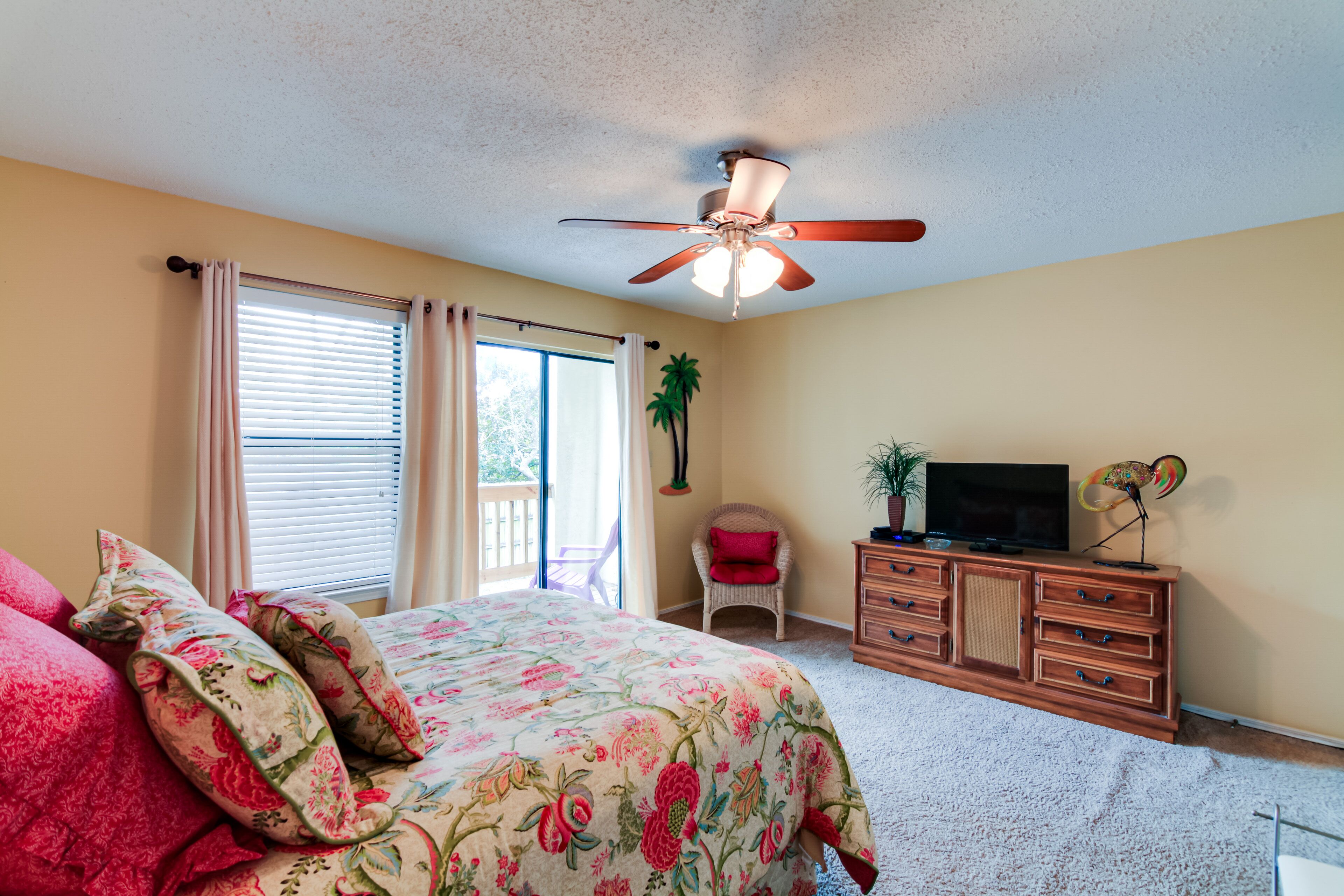 Une chambre à coucher comprenant un lit, un ventilateur de plafond, une commode, un téléviseur et une fenêtre avec des rideaux.