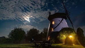 Een groot, cilindervormig gebouw met een kegelvormig dak, omringd door bomen en een picknicktafel in een grasveld.