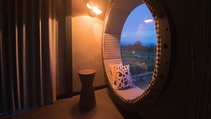 Et værelse med en stribet stol og en lille skammel, et rundt vindue med udsigt, samt en vægmonteret lampe.
