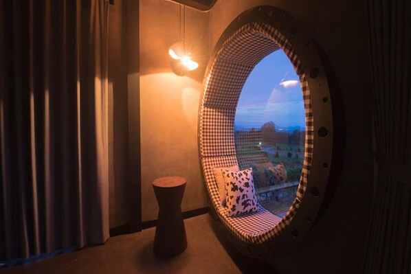 A room with a checkered chair and a small stool, a round window with a view, and a wall-mounted light fixture.