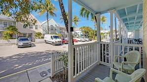 A covered porch with white railings and green chairs, overlooking a street with parked cars and palm trees.