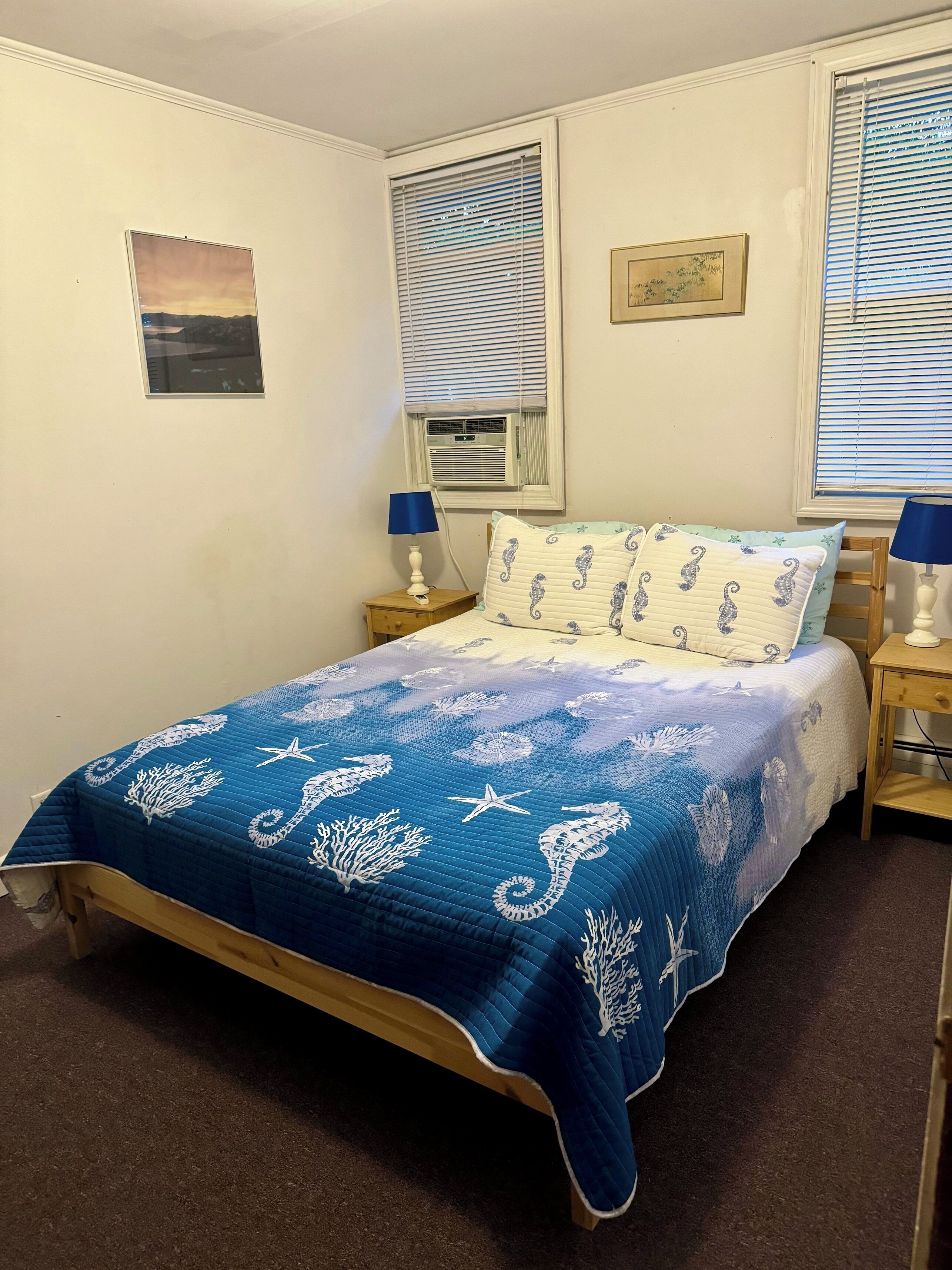 A bedroom with a bed, two nightstands, a window with blinds, an air conditioning unit, and a wall-mounted picture.