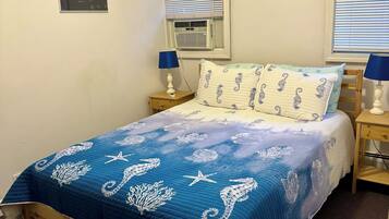 A bedroom with a bed, two nightstands, a window with blinds, an air conditioning unit, and a wall-mounted picture.
