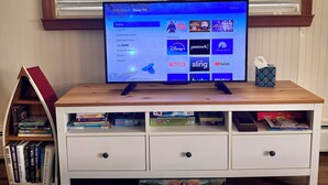 Un televisor sobre un atril de madera con pantalla de menú, una estantería con libros, una caja de pañuelos y un control remoto sobre una mesa.