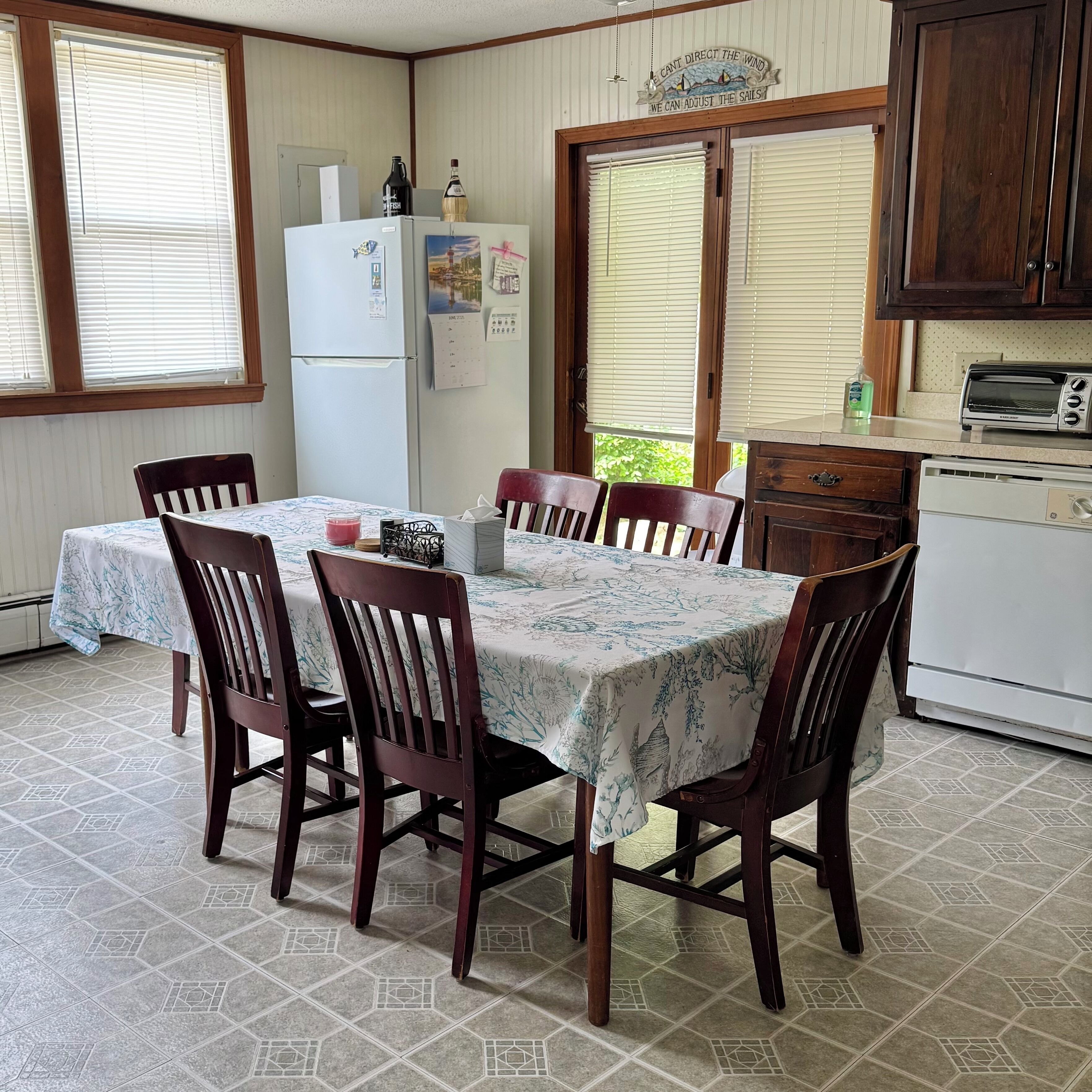 A dining area with a table set for six, surrounded by wooden chairs. A refrigerator and a toaster oven are visible in the background.