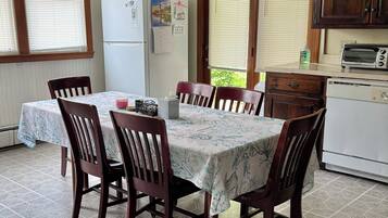 A dining area with a table set for six, surrounded by wooden chairs. A refrigerator and a toaster oven are visible in the background.