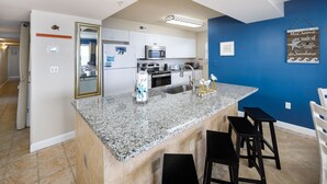 A kitchen with granite countertops, a stainless steel sink, and a microwave. There are three black bar stools and a hanging sign on the wall. - PI 501: Flip Flop Flats - Spectacular East 5th Floor Corner Unit Memory Maker (Fort Walton Beach)