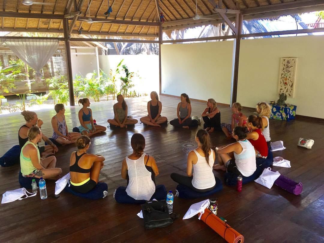 A group of people sitting in a circle on mats in a covered outdoor area with a wooden floor, surrounded by plants and a thatched roof structure.