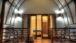 Une pièce avec un plancher en bois, deux fauteuils en osier, un lit recouvert d’une couverture bleue et une porte en bois donnant sur une autre pièce.