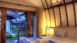 Une chambre à coucher comprenant un lit, une table de chevet, une lampe et donnant sur un espace extérieur doté d’une structure à toit de chaume.