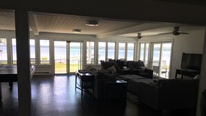A spacious living room with a large window overlooking a body of water, a ceiling fan, and a television. - Modern 300 ft lakefront with pool (Mayville)