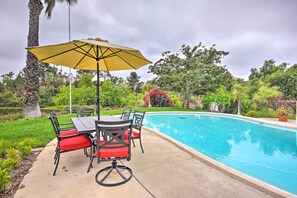 Zona de piscina con mesa y sillas, un paraguas amarillo y una piscina rodeada de vegetación.