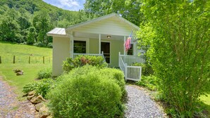 A small, light-colored cabin with a porch and an American flag, surrounded by lush greenery and a gravel path. - Best Getaway for 2 w/ Own Creek & Hot Tub (Marshall)