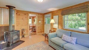 A cozy cabin interior with a wood-burning stove, a sofa, a dining area, and a window with a view of greenery. - Best Getaway for 2 w/ Own Creek & Hot Tub (Marshall)