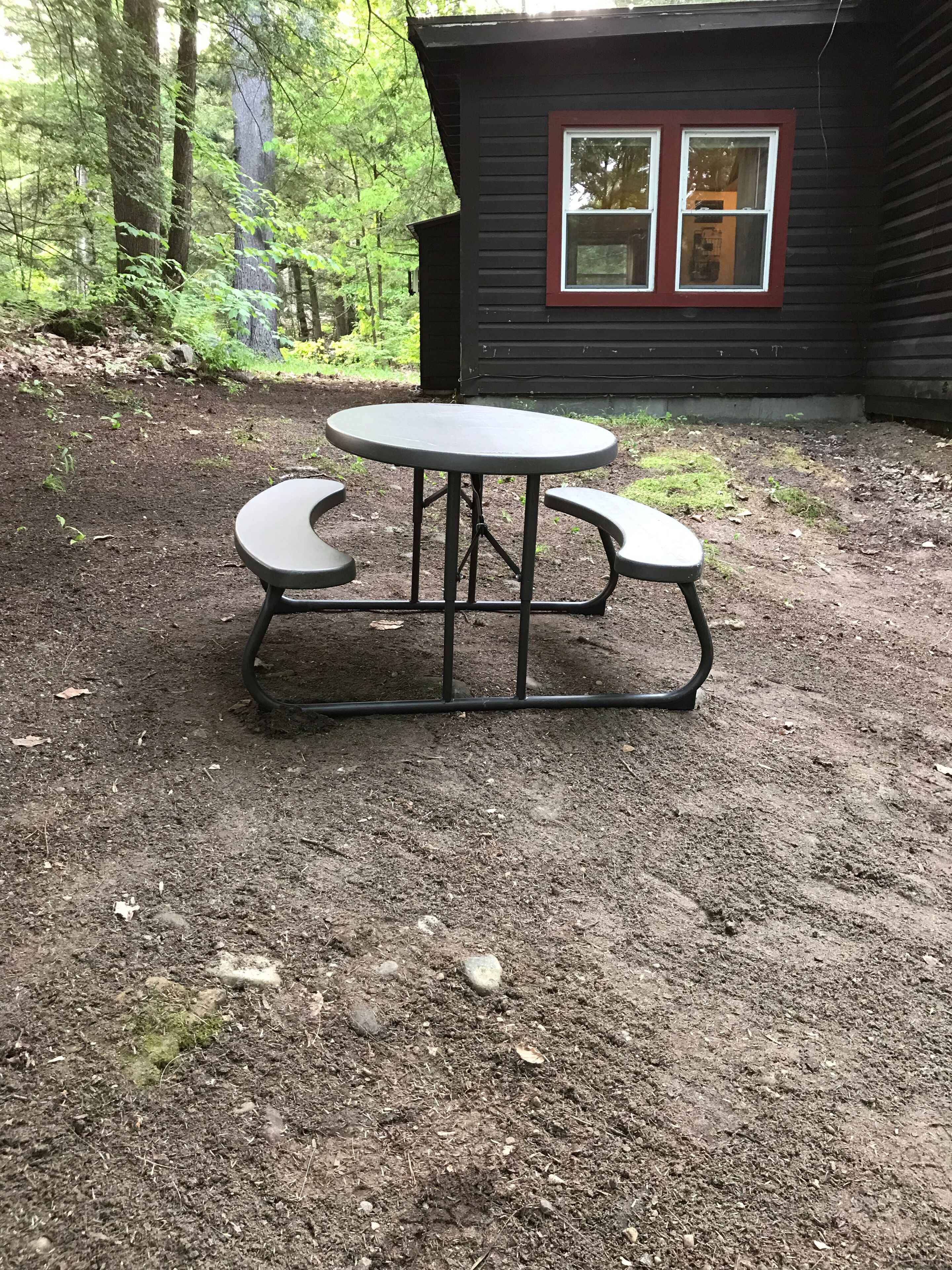 A picnic table with benches in a forested area near a cabin with a red window frame.