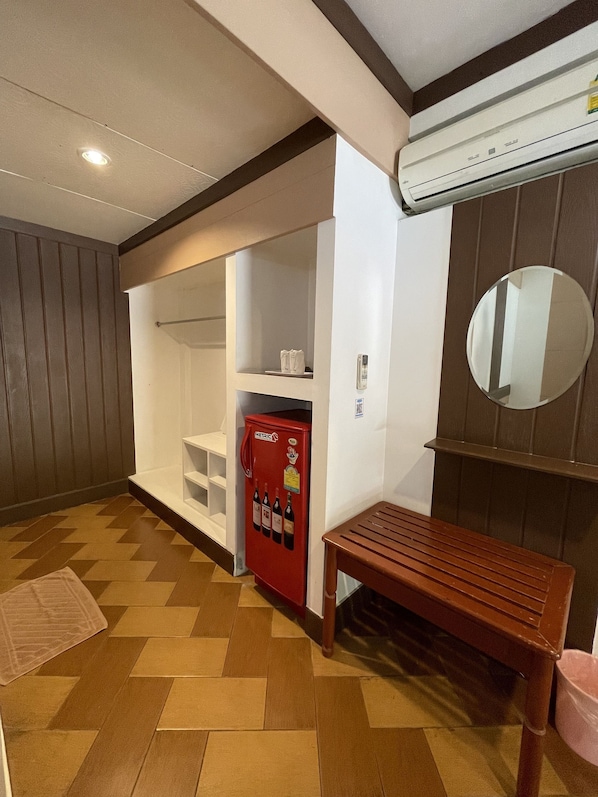 A hotel room with a wooden floor, a red minibar, a white wardrobe, a wooden bench, and a wall-mounted air conditioner. - Bansuan Resort (Nakhon Sawan)