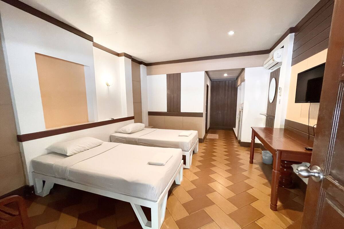 Quarto de hotel com duas camas, uma escrivaninha, televisão e banheiro visível por uma porta.