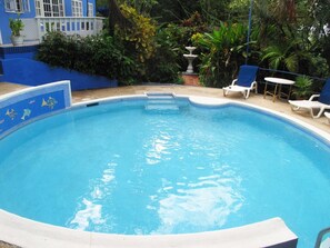 Une piscine dont le bord est recouvert de carreaux bleus, entourée de chaises longues et d’une clôture blanche.