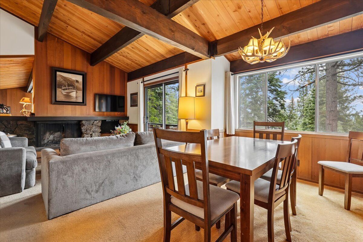 Un salon chaleureux avec une cheminée, des poutres apparentes en bois au plafond, une table à manger avec des chaises et de grandes fenêtres donnant sur la forêt.