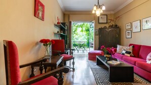 A living room with a red sofa, a wooden coffee table, and a red armchair. There is a bookshelf with books and decorative items, and a window with a view of greenery outside.