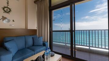 A room with a blue sofa, a small table with a remote control and water bottles, and a large window with a view of the ocean.