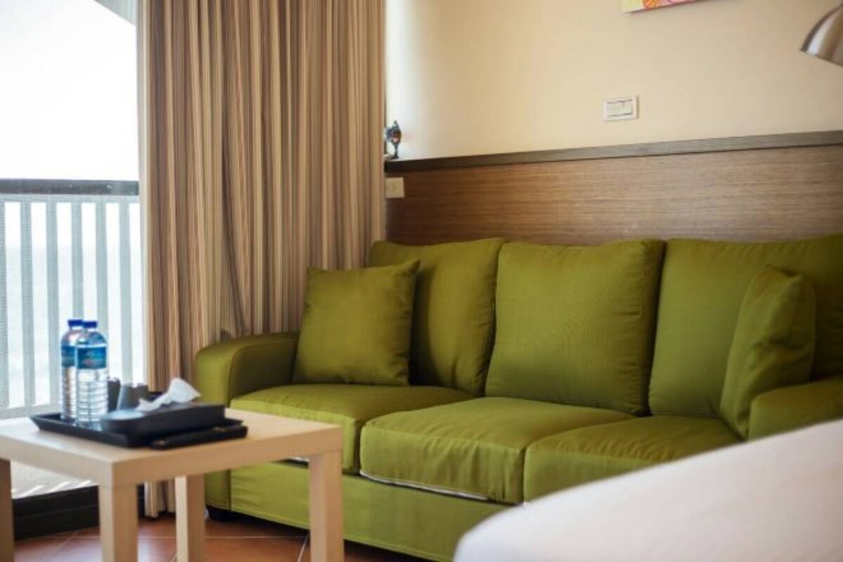 A green sofa with a wooden headboard, a small table with a tray and a water bottle, and a window with curtains.