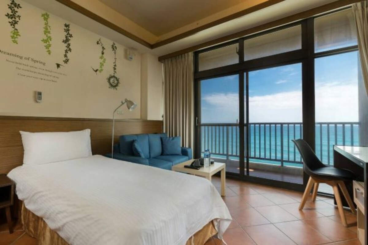 A hotel room with a bed, a blue sofa, a desk, and a view of the ocean through large windows.