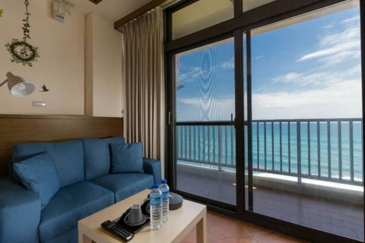 A room with a blue sofa, a small table with a remote control, water bottles, and a view of the ocean through a large sliding glass door.